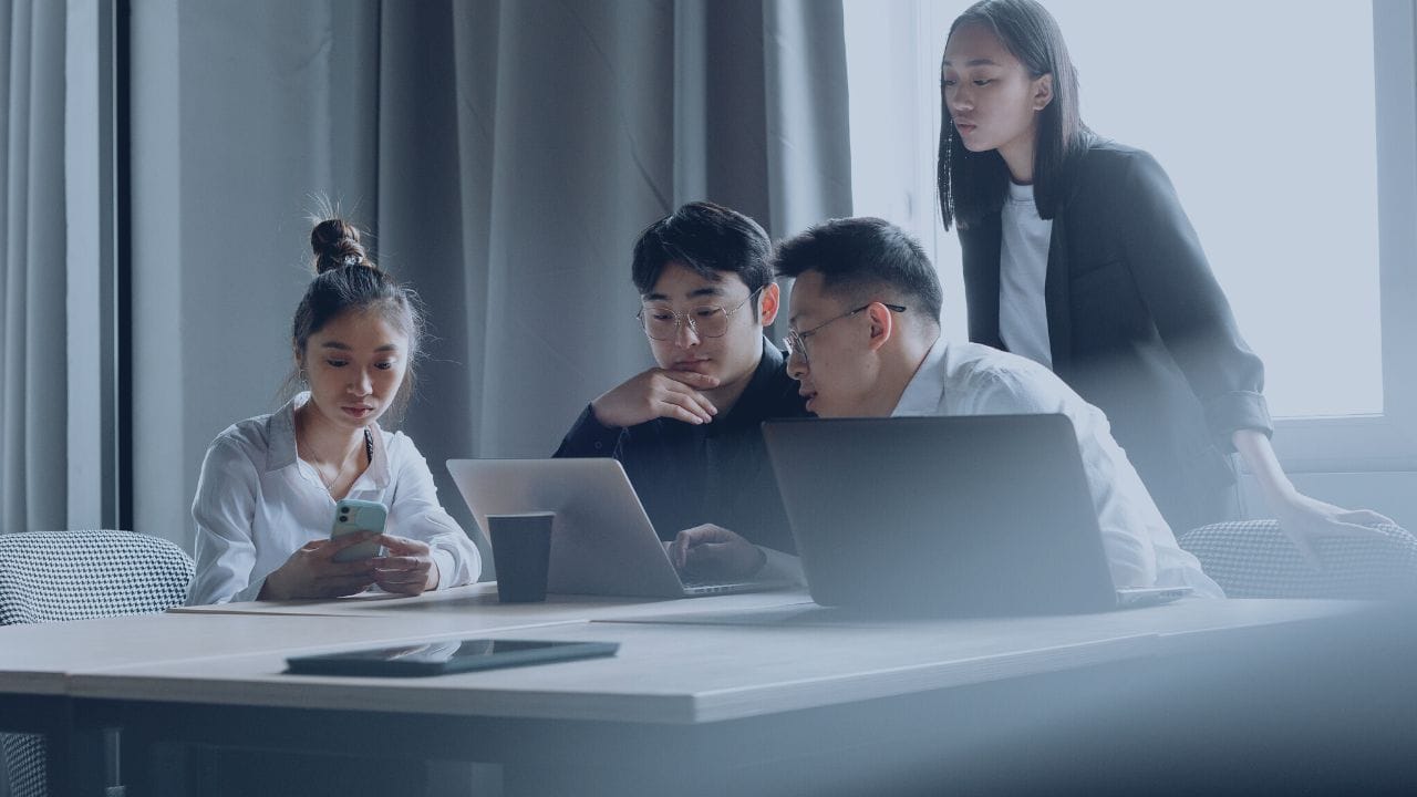 A group of young professionals collaborating in an office, discussing ideas while using laptops and a smartphone during a team meeting.